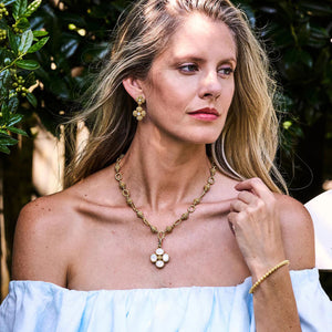 A woman with long blonde hair dons a white off-shoulder top and an 18K gold necklace featuring a pearl pendant. Her accessories include matching earrings and the CAPUCINE DE WULF BERRY ETERNITY PETITE HINGED BANGLE, all set against a backdrop of lush greenery.