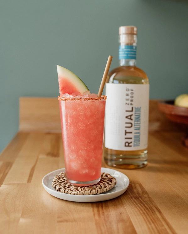 A clear glass bottle of RITUAL ZERO PROOF-TEQUILA ALTERNATIVE by RITUAL ZERO PROOF, a non-alcoholic tequila with a cork top and white label featuring bold black and blue text on a plain background.