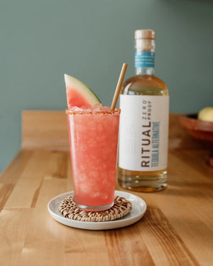 A clear glass bottle of RITUAL ZERO PROOF-TEQUILA ALTERNATIVE by RITUAL ZERO PROOF, a non-alcoholic tequila with a cork top and white label featuring bold black and blue text on a plain background.
