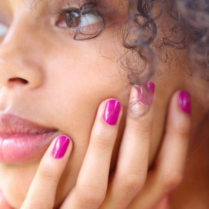 A close-up of MANUCURIST - GREEN™ NAIL POLISH IN ARMERIA by MANUCURIST INC being spread with a brush on a smooth surface, showcasing its vibrant pinky mauve shade, glossy texture, and distinctive curved brushstroke.