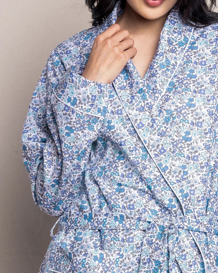 The PETITE PLUME - FLEUR D&#39;AZURE ROBE by PETITE PLUME is a knee-length, long-sleeved bathrobe adorned with a vintage-inspired floral print in blue and white hues. Crafted from the finest quality cotton, the robe features a shawl collar and includes a matching fabric belt tied at the waist. It offers a relaxed fit ideal for comfortable lounging in luxurious sleepwear.