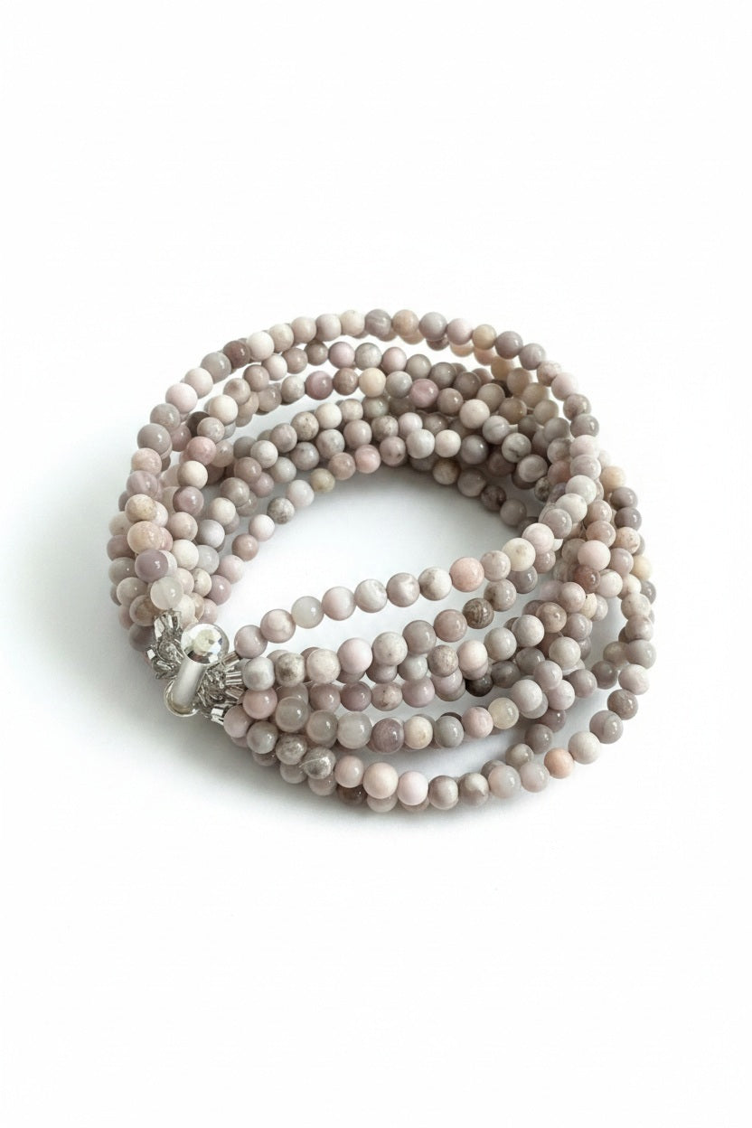 The ANN LIGHTFOOT - NINE STRAND SAKURA AGATE CUFF features smooth, round sakura agate beads in light grey and beige. Coiled with multi-strands, the bracelet has 4mm beads and is secured by a stainless steel clasp against a plain white backdrop.