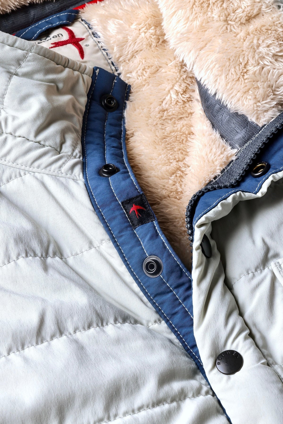 The RELWEN Quilted Superfleece Popover in Chalk is a beige fleece jacket with quilted green sleeves and white stripes, shown laid flat on a white background. This semi-water resistant fall-winter coat features a high collar and stylish navy blue trim along the zipper and collar for added protection and flair.