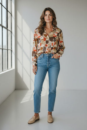 A woman with wavy brown hair stands barefoot indoors, wearing brown wide-leg pants and the HARTFORD HATFORD - CINEMA SHIRT GEOMETRIC PRINT—a loose-fit shirt featuring an abstract orange, beige, and gray pattern—as she gazes out a large window.