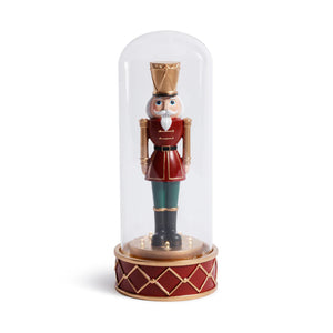 The SOLDIER IN CLOCHE by ZODAX is a decorative nutcracker with a white beard, red uniform, green pants, and gold accents, standing upright inside a clear glass dome on a red and gold base; battery operated for convenience.