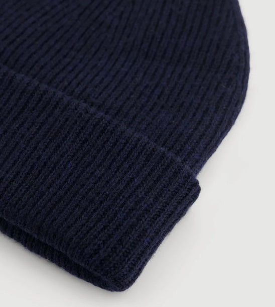 The HARTFORD - SEAMLESS WOOL BEANIE IN NAVY by HARTFORD is a dark blue ribbed knit beanie made from 100% wool, featuring a folded brim and shown against a plain light gray background.