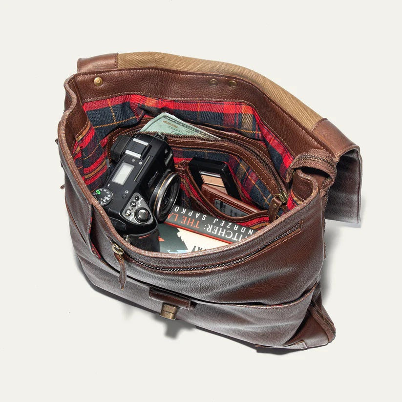 The WILL LEATHER GOODS - OTTO CROSSBODY BAG IN COGNAC, a full-grain leather bag with plaid lining, is shown open from above holding a camera, book, and wallet.