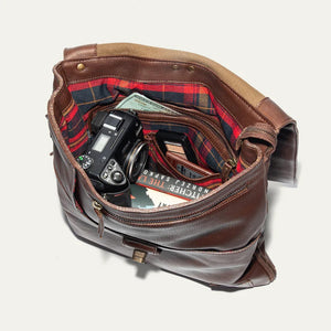 The WILL LEATHER GOODS - OTTO CROSSBODY BAG IN COGNAC, a full-grain leather bag with plaid lining, is shown open from above holding a camera, book, and wallet.