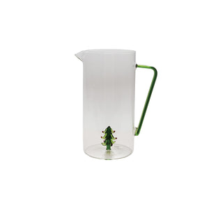 A CREATIVE COOP GLASS PITCHER WITH HOLIDAY TREE sits on a wooden surface with a green-stemmed glass holding snowy holiday trees beside it, set against a light background.