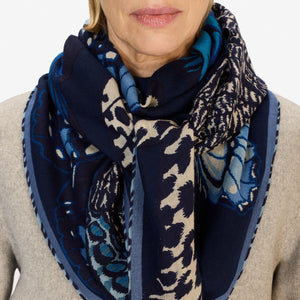 A woman with light hair pulled back wears a light gray jacket and the INOUI EDITIONS PAPILLON (BUTTERFLIES) square wool scarf in blue by INOUI. She stands against a plain white background, looking straight at the camera with her hands in her pockets.
