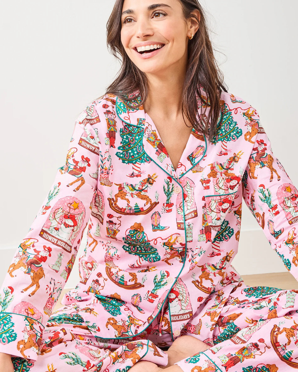 A woman sits on the floor against a plain white wall, wearing the PRINTFRESH - COWBOY CHRISTMAS LONG PAJAMA SET by PRINTFRESH, featuring pink fabric with festive Santa, reindeer, wreath prints, and "Howdy Holidays" text.