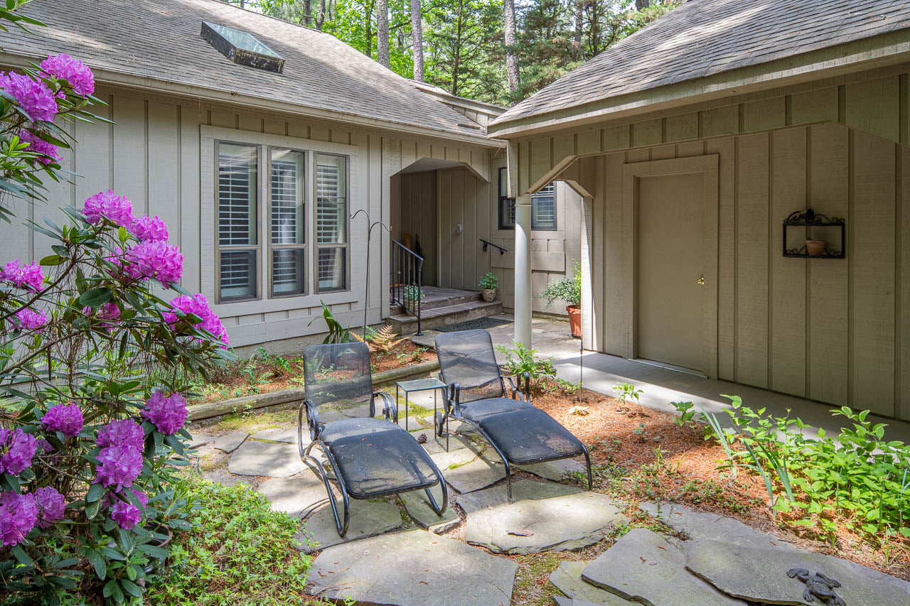 A shaded garden courtyard with three black lounge chairs on stone pavers, surrounded by blooming pink flowers and greenery, next to a beige house with large windows and a covered walkway.