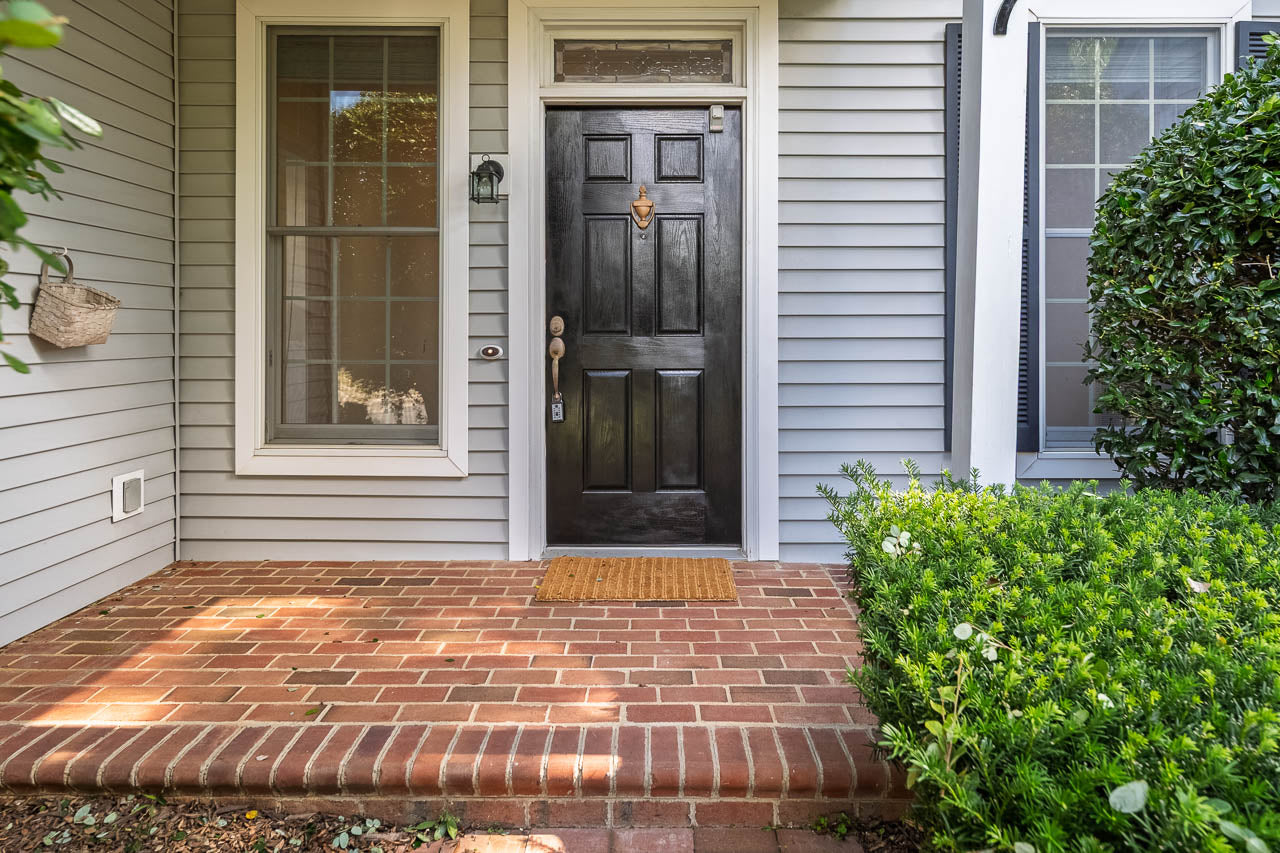 A black front door with a brass knocker and handle is centered between two tall windows on a gray house with white trim. A tan doormat sits on a red brick porch surrounded by green bushes.