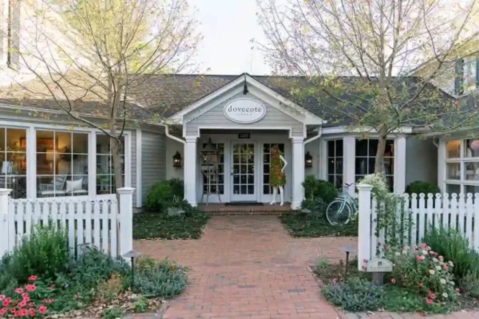 A charming white cottage with large windows, a central entrance, white picket fence, leafy garden, and bicycle parked out front. A sign above the door reads “dovecote.”.