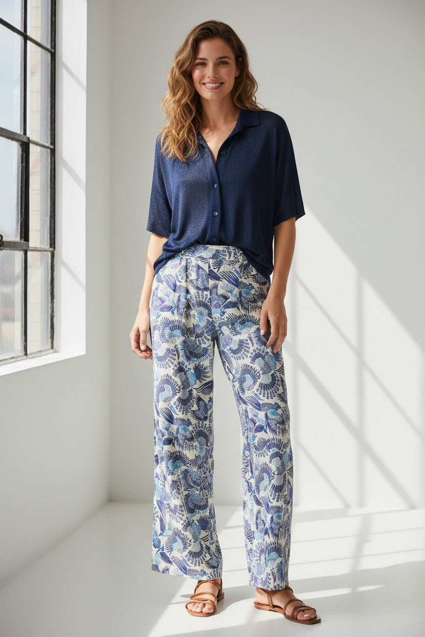 The MOMONI - ASPEN PANT features a luxurious haboutai silk fabric with a geometric pattern of squares and rectangles, adorned with vibrant floral motifs and architectural designs in colors like yellow, green, and blue.