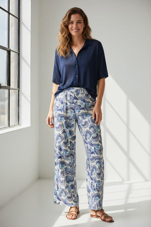 The MOMONI - ASPEN PANT features a luxurious haboutai silk fabric with a geometric pattern of squares and rectangles, adorned with vibrant floral motifs and architectural designs in colors like yellow, green, and blue.