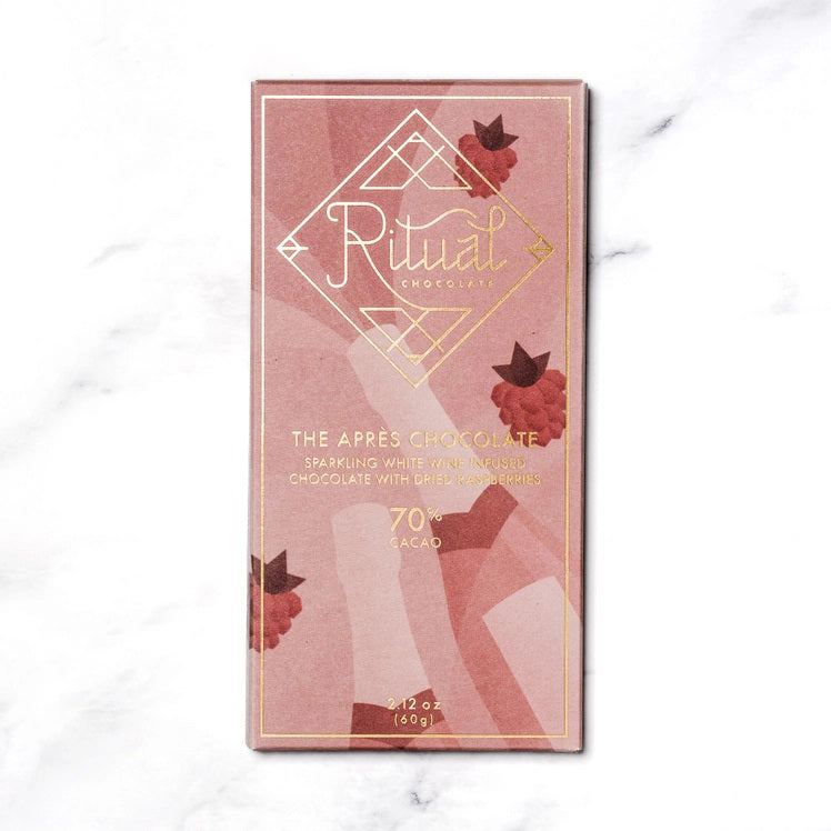 A rectangular chocolate bar with a pinkish wrapper adorned with minimalist illustrations of raspberries and abstract shapes. The packaging reads "RITUAL CHOCOLATE - THE APRES CHOCOLATE BAR 70%, Sparkling Wine Infused Chocolate with Dried Raspberries," 2.12 oz (60g).