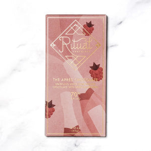 A rectangular chocolate bar with a pinkish wrapper adorned with minimalist illustrations of raspberries and abstract shapes. The packaging reads "RITUAL CHOCOLATE - THE APRES CHOCOLATE BAR 70%, Sparkling Wine Infused Chocolate with Dried Raspberries," 2.12 oz (60g).