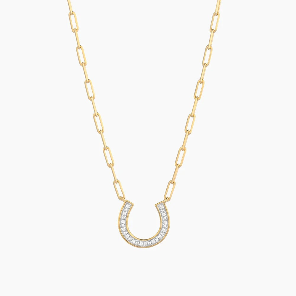 A woman wearing the ELLA STEIN - LUCKY HORSE SHOE PENDANT NECKLACE, crafted from gold plated stainless steel and adorned with natural diamonds, stands outdoors in front of a blurred beach background. This elegant, waterproof piece by ELLA STEIN is perfect for effortless seaside style.