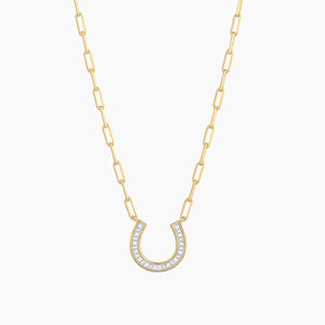 A woman wearing the ELLA STEIN - LUCKY HORSE SHOE PENDANT NECKLACE, crafted from gold plated stainless steel and adorned with natural diamonds, stands outdoors in front of a blurred beach background. This elegant, waterproof piece by ELLA STEIN is perfect for effortless seaside style.