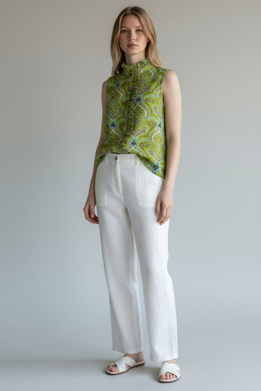 A woman with light hair stands calmly against a plain background, wearing the HOPE FOR FLOWERS FOPE FOR FLOWERS sleeveless frilled button-down in green foulard, paired with white high-waisted pants and white sandals. Her arms rest relaxed at her sides.