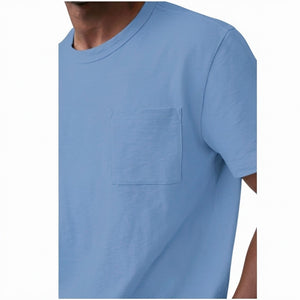 A man with short curly hair wears the MACK WELDON Heavyweight Slub Cotton Pocket T-Shirt in Dean Blue with dark pants, standing against a plain white background.