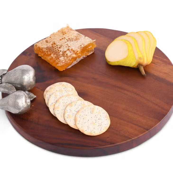 The VAGABOND HOUSE PEAR CHEESE TRAY is a round wooden cutting board with a smooth surface made from richly colored wood. It features two small decorative pears resembling pure-pewter designs on the right side, set against a plain white background, highlighting its elegance.