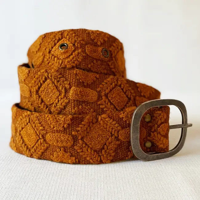 The JENNY KRAUSS - TERRACOTTA EMBROIDERED WOOL BELT, handcrafted in Peru, features a diamond embroidery pattern, metal buckle, and textured brown wool fabric, neatly coiled on a white surface.