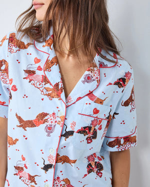 A woman with long, wavy brown hair stands barefoot against a white wall, wearing the PRINTFRESH Dachshund Date Short Sleeve Top & Cropped Pants Pajama Set in Blue. The organic cotton set from PRINTFRESH features short sleeves and cropped pants adorned with a charming dachshund print. She smiles slightly.