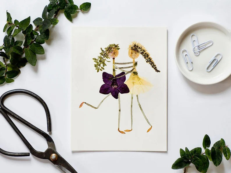 The PETAL PEOPLE PRESS - BEST FRIENDS CARD by PETAL PEOPLE PRESS features two whimsical figures crafted from dried flowers and leaves. Against a white background, one figure dons a purple flower and the other a yellow one, creating a playful, nature-inspired scene ideal for any greeting card.