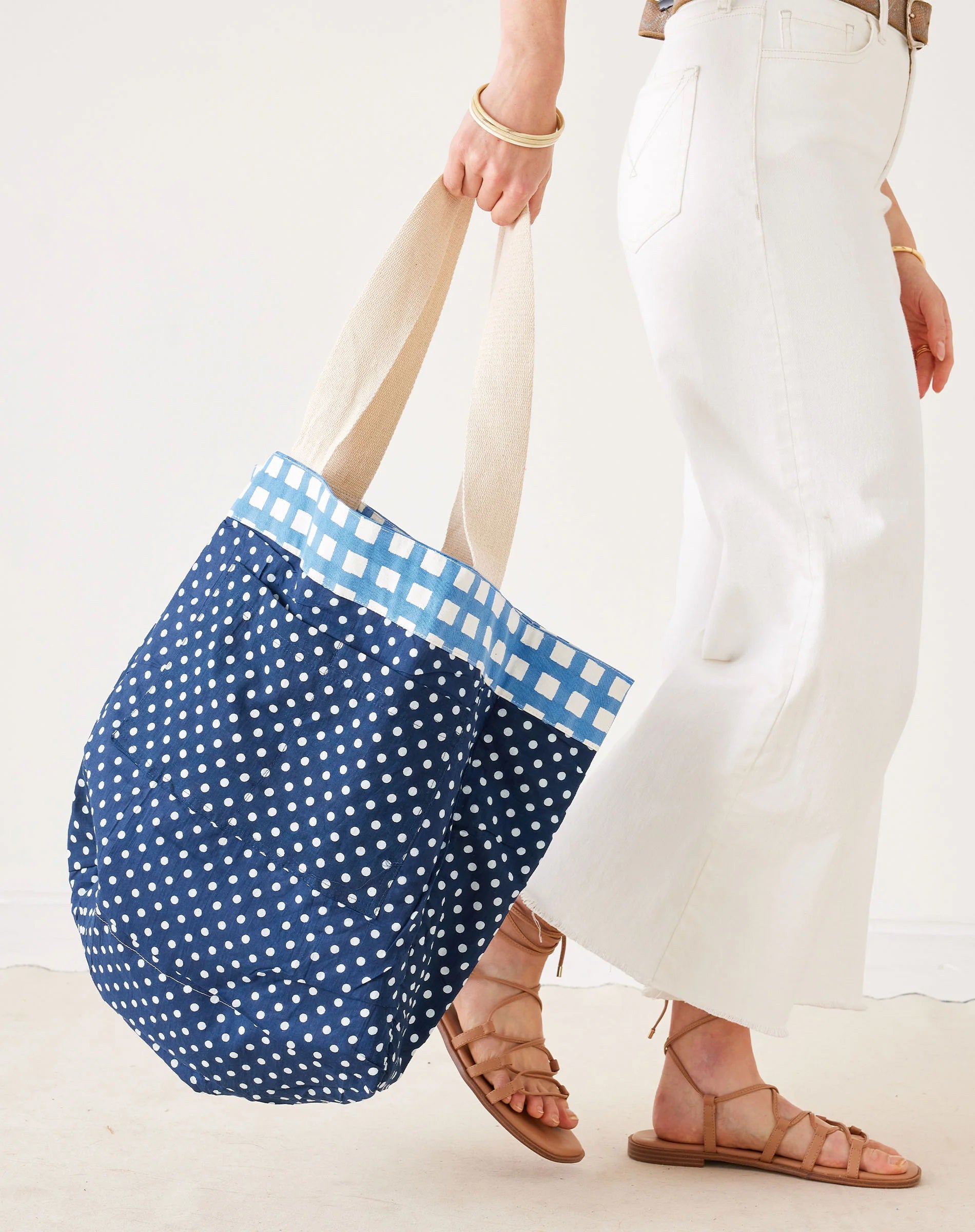 A person wearing white pants and brown sandals carries a large blue polka dot tote bag with cream handles and a blue gingham trim.