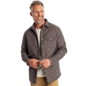 A smiling man with gray hair and a beard wears the CRIQUET - QUILTED SHACKET IN CHARCOAL by CRIQUET over a light shirt and tan pants, standing against a white background.