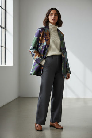 A woman in a white turtleneck and black pants stands against a plain background, layering her look with the ALYKI LABORATORIO PRINTED SATIN JACKET, featuring bold, colorful green, purple, brown, and blue leaf patterns.