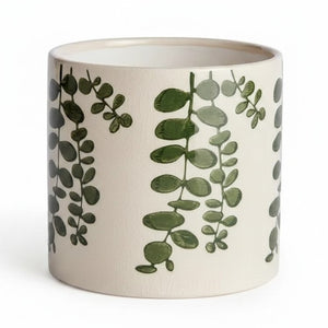 Two NAPA HOME AND GARDEN Maren Pots in white ceramic with an embossed leaf pattern sit on a wooden table, one with a leafy plant. Nearby are two stacked books and a partially visible empty green cachepot.