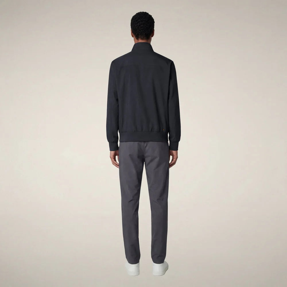 A person stands facing forward, wearing the SAVE THE DUCK - TULIO JACKET IN BLUE BLACK by SAVE THE DUCK, paired with gray pants and white sneakers, against a plain light background.