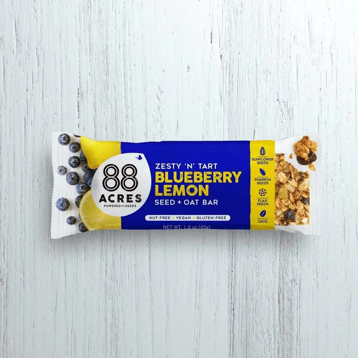 A zesty &#39;N&#39; tart 88 ACRES - BLUEBERRY LEMON SEED OAT BAR is displayed against a light wooden surface. The blue and white packaging showcases images of blueberries and lemon. This bar from 88 ACRES is nut-free, vegan, gluten-free, and made with sunflower seeds.