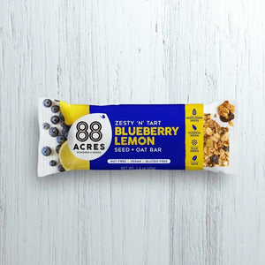 A zesty 'N' tart 88 ACRES - BLUEBERRY LEMON SEED OAT BAR is displayed against a light wooden surface. The blue and white packaging showcases images of blueberries and lemon. This bar from 88 ACRES is nut-free, vegan, gluten-free, and made with sunflower seeds.