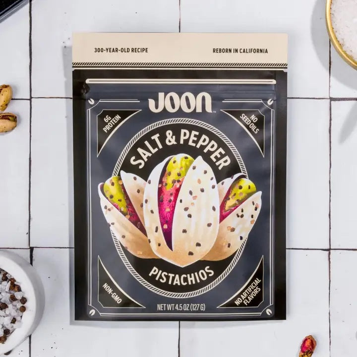 A bag of JOON- SALT & PEPPER PISTACHIOS by JOON, a savory plant-based snack, rests on a white tiled surface with pistachios and a bowl of salt nearby. The packaging features claims such as non-GMO, no artificial flavors, and 6g of protein per serving.