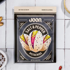 A bag of JOON- SALT & PEPPER PISTACHIOS by JOON, a savory plant-based snack, rests on a white tiled surface with pistachios and a bowl of salt nearby. The packaging features claims such as non-GMO, no artificial flavors, and 6g of protein per serving.