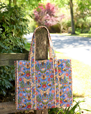 The PRINTFRESH - Bee Tapestry Traveler Tote in Delphinium Blue by PRINTFRESH, featuring vibrant floral, butterfly, and bee designs on organic cotton, hangs on a rustic wooden fence outdoors with lush green foliage in the background.