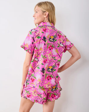 A woman wearing the PRINTFRESH Bird & Blossom Ruffle Pajama Short Set, featuring a pink, nature-inspired floral and bird print, stands against a plain light-colored wall with one hand in her shorts pocket, looking at the camera.