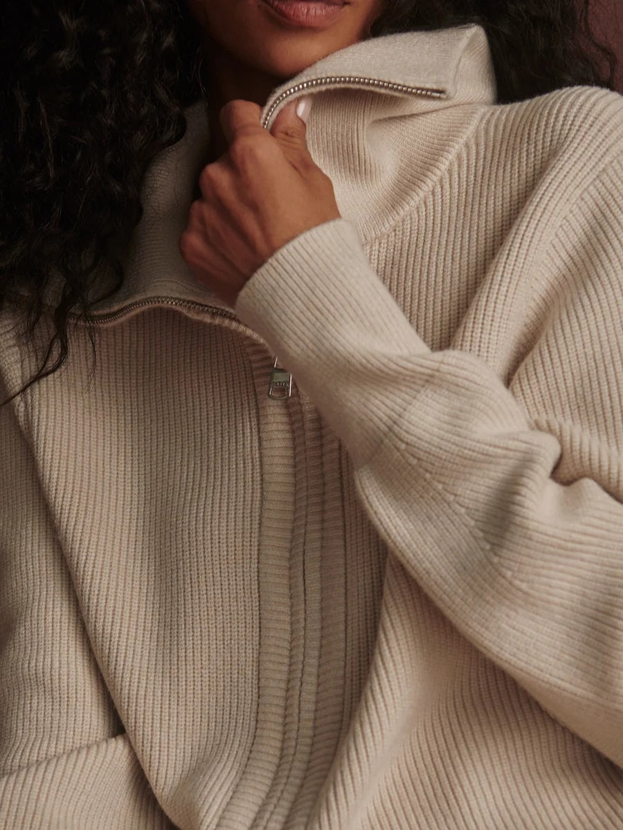 A person wearing a dark brown VARLEY - ENRICO ZIP-THROUGH KNIT JACKET featuring a ribbed soft viscose knit, a high collar, and a front zipper, ideal for cool weather layering.