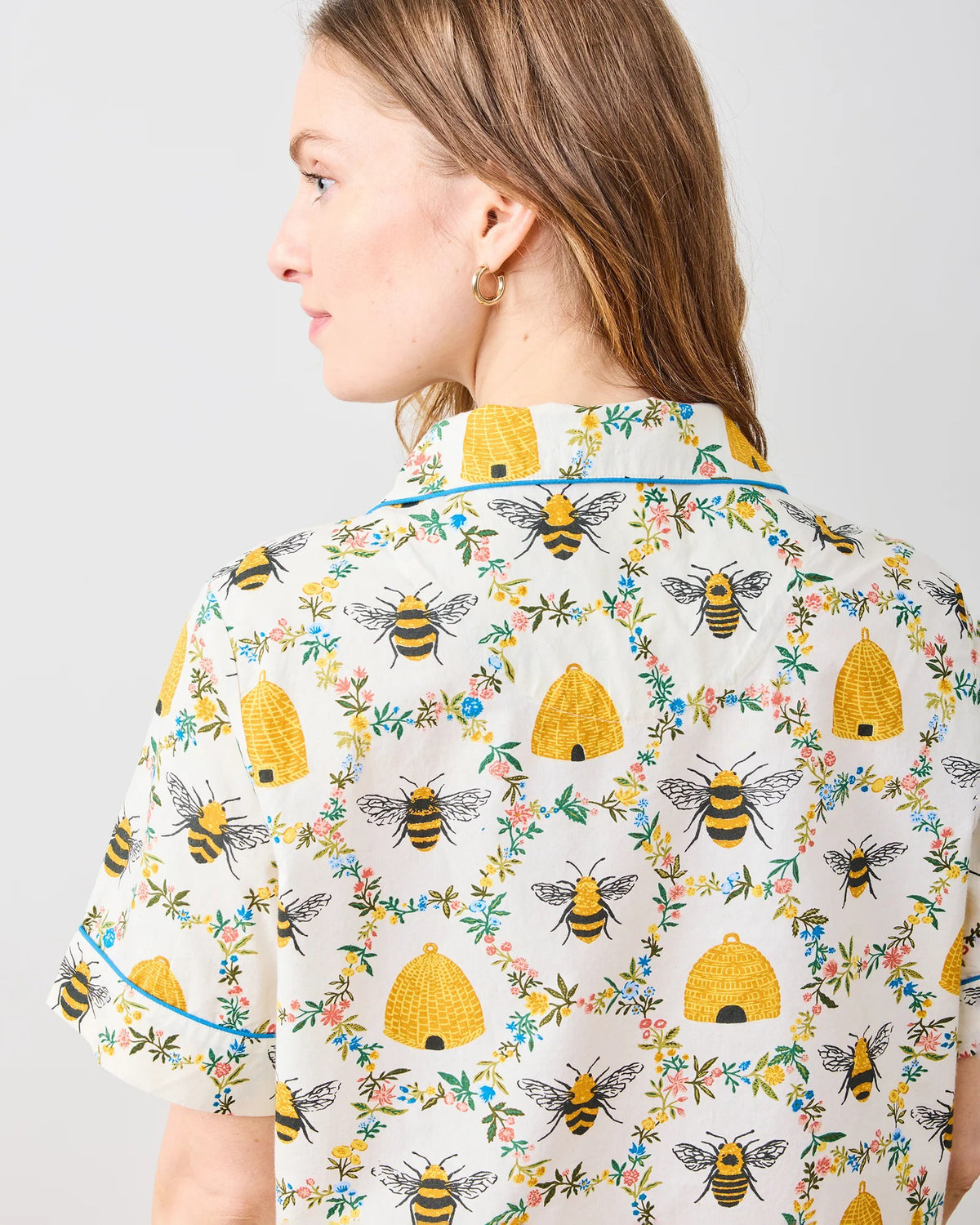 A person with long hair is wearing the PRINTFRESH - SWEET BEE COTTON SHORT SET, featuring a floral pattern in yellow and green. The short-sleeved shirt and shorts exude comfort and style as they stand barefoot on a wooden floor against a plain white wall.