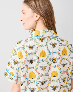 A person with long hair is wearing the PRINTFRESH - SWEET BEE COTTON SHORT SET, featuring a floral pattern in yellow and green. The short-sleeved shirt and shorts exude comfort and style as they stand barefoot on a wooden floor against a plain white wall.