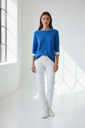A woman with straight brown hair and gold earrings wears the HINSON WU TYLER 3/4 SLEEVE SWEATER in bright blue, styled with rolled cuffs, white pants, and a gold bracelet, standing against a neutral background.