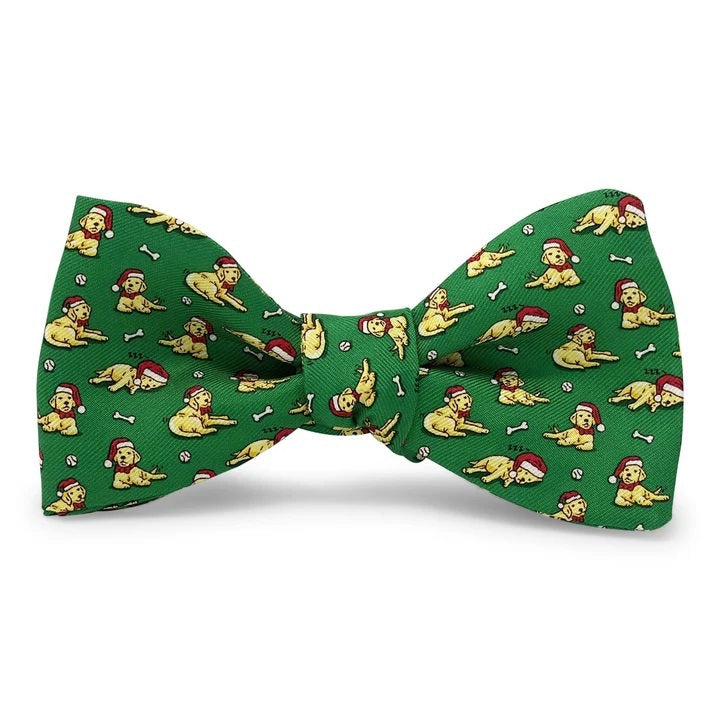 Add a touch of holiday cheer to your ensemble with the BIRD DOG BAY - SANTA PAWS SILK BOW TIE IN MID-GREEN. This playful, handmade bow tie by BIRD DOG BAY features a mid-green silk background adorned with a repeating pattern of yellow dogs wearing red hats, chewing on bones, and playing with balls—making it the perfect festive accessory for any holiday gathering.