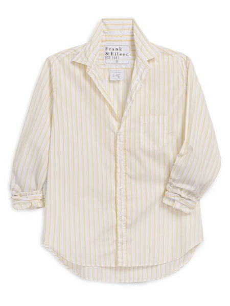 A woman with wavy brown hair smiles slightly, wearing the FRANK &amp; EILEEN EILEEN Relaxed Button Up Shirt in Italian Cotton Thin Yellow Stripe, paired with blue jeans and white sneakers. She stands in a plain, well-lit background.