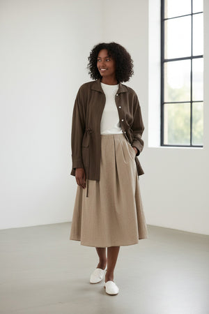 Someone wears the ELEH - JACKET WITH TIE WAIST by ELEH over a white t-shirt and a light beige textured skirt, standing against a plain light background.