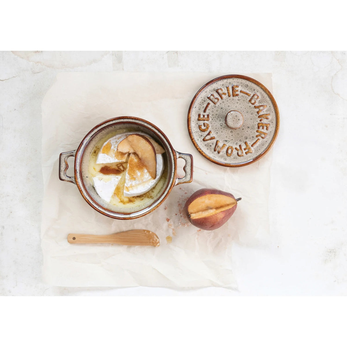 A Brie cheese sits in the CREATIVE COOP STONEWARE BRIE BAKER WITH LID AND BAMBOO SPREADER, topped with pear and honey. The lid is set aside, while a bamboo spreader and half a pear rest nearby on parchment paper.
