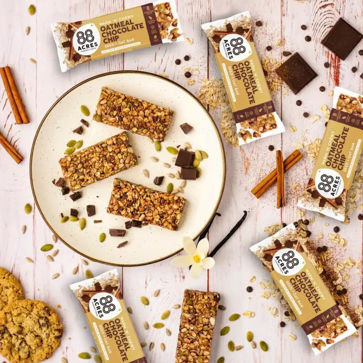 A packaged snack bar labeled &quot;88 ACRES - OATMEAL CHOCOLATE CHIP SEED OAT BAR&quot; from the brand 88 ACRES is shown against a light wooden background. The packaging highlights that it is a seed + oat bar, emphasizing its nut-free, vegan, and gluten-free qualities, and promises creamy chocolate chips for a delightful chewy texture.